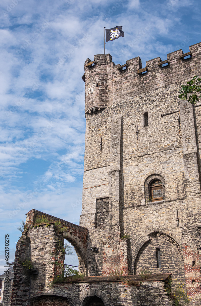 Gent, Flanders, Belgium - July 30, 2021: South-East corner of gray-brown wall and tower of medieval Gravensteen stone castle under blue cloudscape with some green foliage and flag.