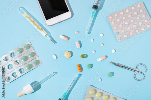 Tablets, pills, scissors, pipette, plastic medical disposable syringes, clinical manual thermometer and white phone on pastel blue background. Minimal creative medical equipment concept. 