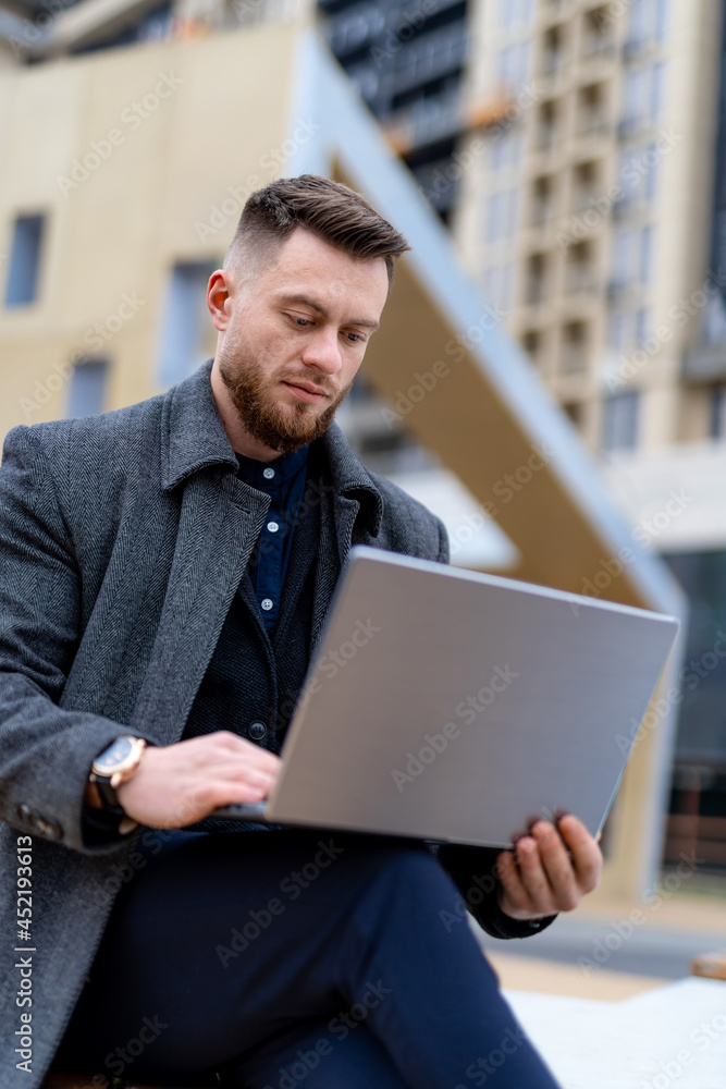 Persistent businessman in gray casual coat doing work on laptop. Modern wide street on background. Business concept Persistent businessman in gray casual coat doing work on laptop. Modern wide street on background. Business concept