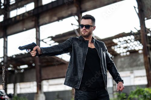 Man with a beard in a black leather jacket, wearing sunglasses and holding a gun. Man is aiming somewhere. Guns concept photo