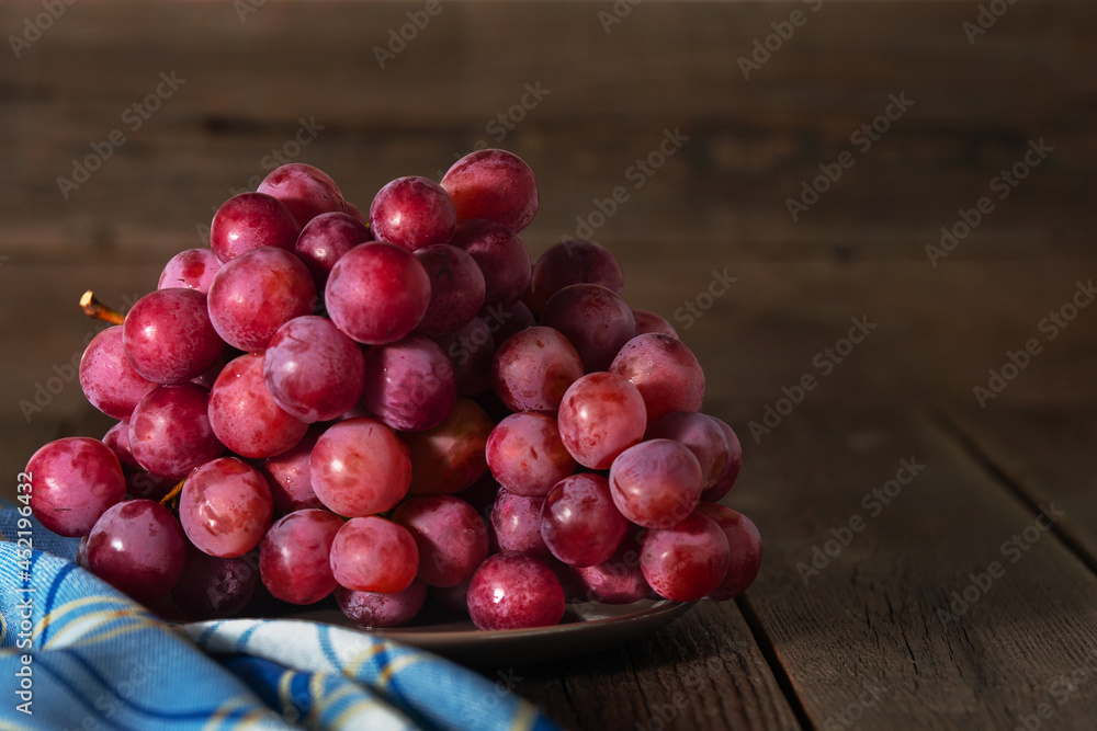 On a wooden background in a plate lies a bunch of red ripe grapes. In the foreground is a checkered napkin. On a wooden background in a plate lies a bunch of red ripe grapes. In the foreground is a checkered napkin.