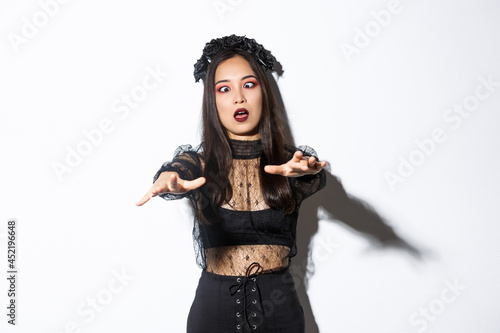 Image of funny and beautiful woman acting like undead widow, wearing black lace dress and wreat, looking like zombie and moving forward. Female enjoying halloween party
