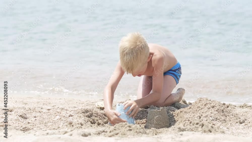 Little blonde boy playing with sand on beach ocean sea. Child building sand castle house. Family summer holidays and trips to warm countries Little blonde boy playing with sand on beach ocean sea. Child building sand castle house. Family summer holidays and trips to warm countries