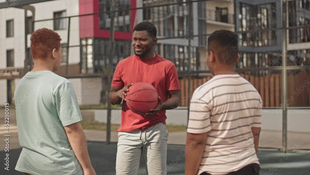 Medium shot of joyful African American man teaching his two teenage sons to play basketball practicing outdoors at sportsground on sunny day Medium shot of joyful African American man teaching his two teenage sons to play basketball practicing outdoors at sportsground on sunny day