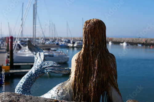 Brixham, Devon, England - May. 30. 2021: Mermaid sculpture sitting overlooking marina and the Brixham lifeboat - 'Duke of Kent'. photo