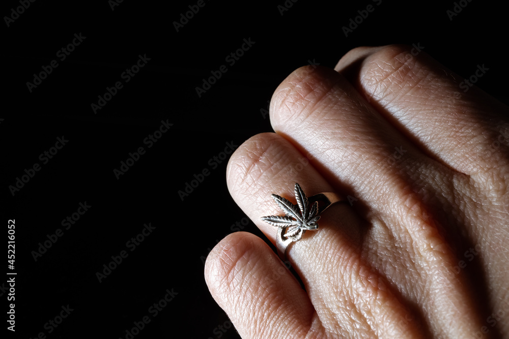 Obraz premium Close-up of a silver ring in the form of a marijuana leaf worn on a hand. White gold jewelry. Drug addiction symbol. Illegal distribution of harmful substances. Sign. Banner. Copy space. Dealer sign