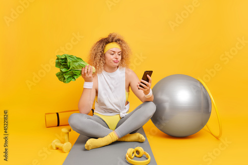 Full length shot of curly haired woman sits crosses legs on fitness mat holds green vegetable keeps to diet checks how much calories she burnt after workout via smartphone application trains indoor
