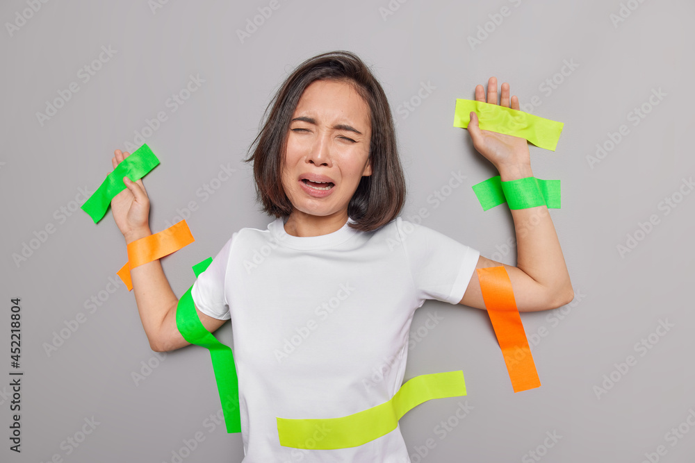 Upset crying Asian woman with dark hair feels discontent being victim of abusement stuck with colorful plasters to wall wears casual t shirt isolated over grey background. Female model in captivity Upset crying Asian woman with dark hair feels discontent being victim of abusement stuck with colorful plasters to wall wears casual t shirt isolated over grey background. Female model in captivity