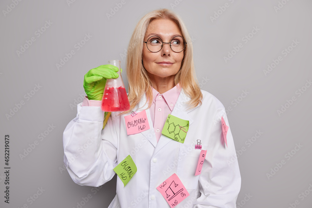 Serious professional female chemist wears round spectacles white coat with stuck stickers written formulas holds glass flask with red liquid after mixing reagents isolated over grey background Serious professional female chemist wears round spectacles white coat with stuck stickers written formulas holds glass flask with red liquid after mixing reagents isolated over grey background