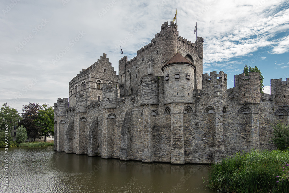 Gent, Flanders, Belgium - July 30, 2021: West side of gray Gravensteem medieval stone castle under blue cloudscape behind dark water moat. Some green foliage.