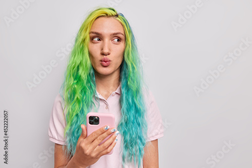 Beautiful teenage girl with trendy colorful dyed hair keeps lips folded looks away dressed in casual t shirt uses mobile phone for chatting online isolated over grey background blank copy space