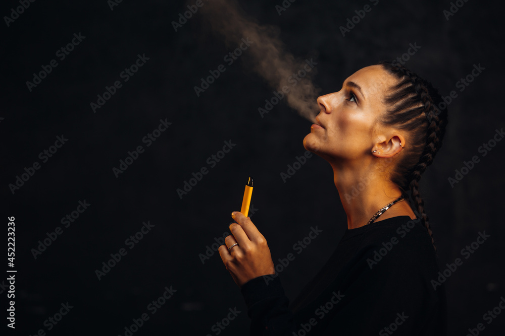 Fototapeta premium Beautiful young woman in black background with backlight smokes electronic cigarette