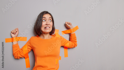 Frightened Asian woman with dark hair stuck with adhesive tapes to grey wall bites lips has nervous expression isolated over grey background blank space for your advertisement. Captured female