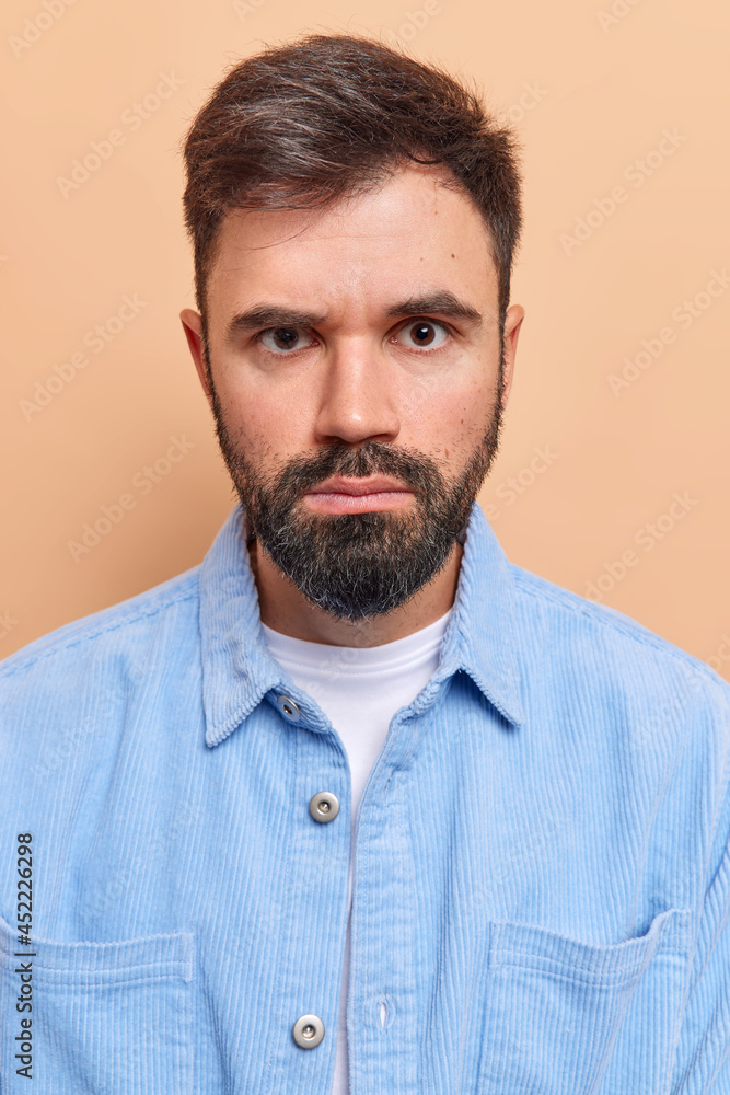 Obraz premium Vertical shot of handsome bearded man looks seriously has strict expression gazes directly at camera wears blue velvet shirt smirks face isolated over beige background. Human face expressions