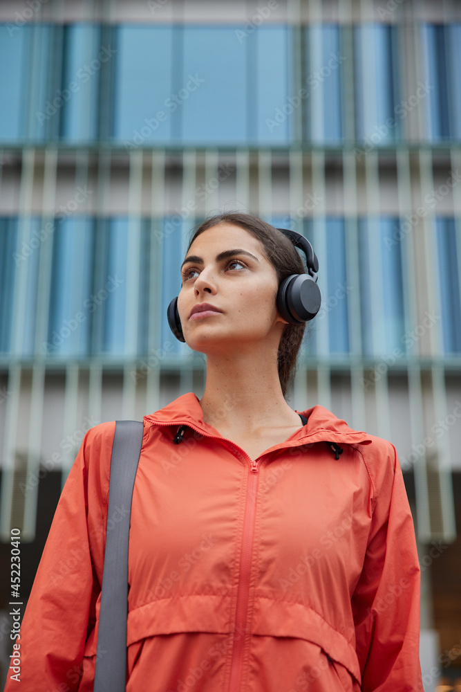 Φωτογραφία Vertical shot of thoughtful brunette woman listens music via headphons enjoys favorite playlist wears anorak strolls in downtown poses against modern building