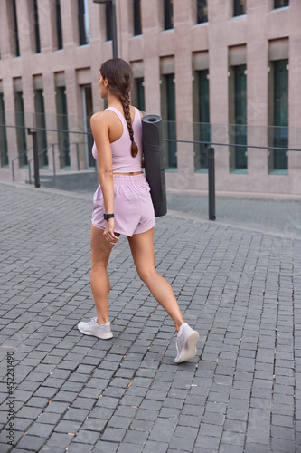 Back view of slim sporty woman in activewear walks outdoors carries fitness mat on shoulder returns from training poses in center of city near modern buildings. Sportswoman looks for place for workout © wayhome.studio 