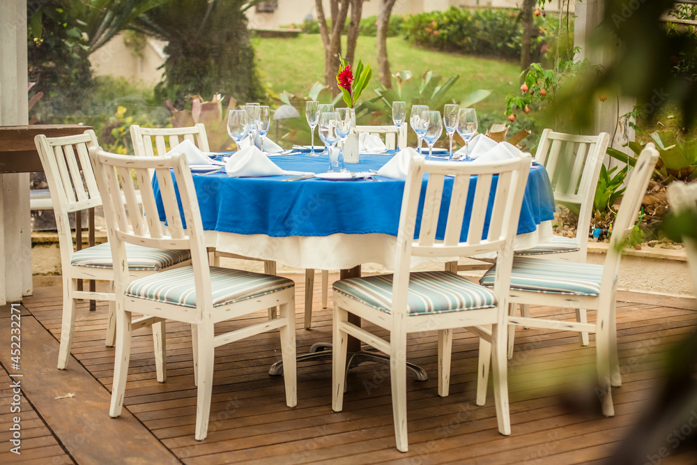 Obraz premium Outdoor restaurant table during the day with chairs, tableware, napkins and a red flower in the center - Corporate lunch or wedding party with garden in the back