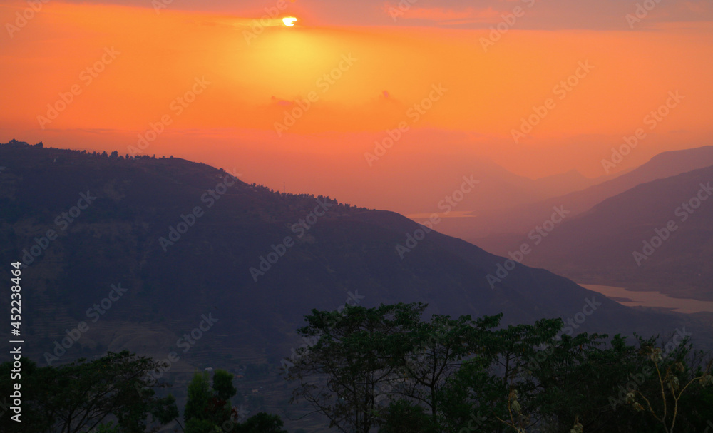 A sunset with deep orange saffron  sky over the mountains crossing each other and valley overlooking the trees