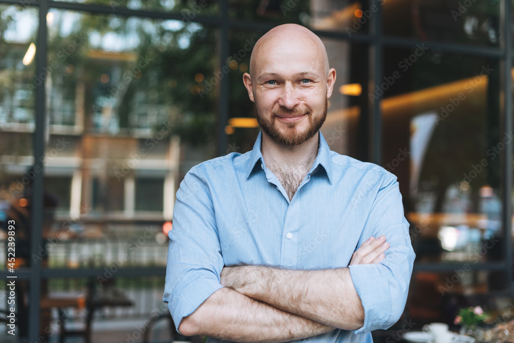 Portrait of Adult bald smiling attractive man forty years with beard in blue shirt businessman against glass wall of the street cafe - obrazy, fototapety, plakaty 