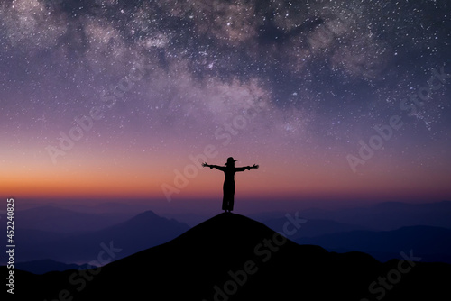 Silhouette of young female traveler wearing hat standing and open arms watched the beautiful night sky, star and milky way alone on top of the mountain.