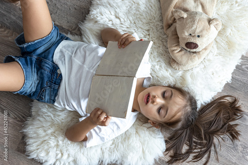 Slika na platnu Little girl reads a book lying on the floor at home, top view.