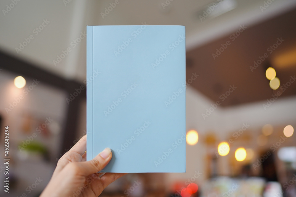 Woman hand holding a light blue book with blank cover for inserting text on blurred cafe background. Woman hand holding a light blue book with blank cover for inserting text on blurred cafe background.