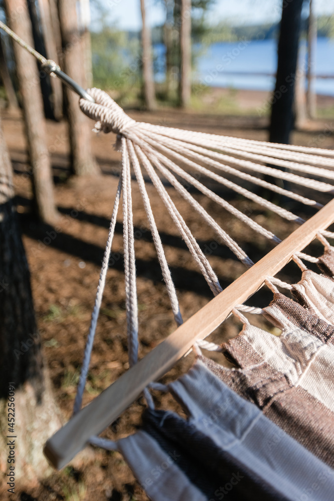A neutral-colored hammock with a fringe hangs between the pines on the river bank. Close-up of the details. the concept of a picnic and outdoor recreation on a sunny summer day.