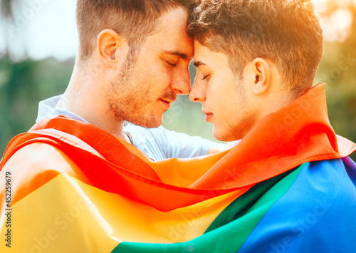 Gay couple with colourful LGBT flag enjoying nature outdoors, kissing and hugging. Young men family in love concept. Happiness
