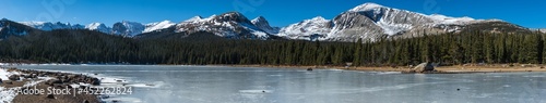 A wide angle panoramic landscape shot of snowy mountains, a line of pine trees, and a frozen lake and shore line panorama