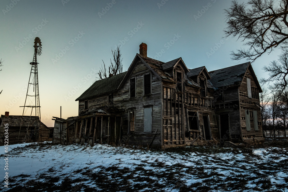 A wide angle landscape shot of a creepy, abandoned house made of wooden planks in the snow with a windmill and trees shot during the winter