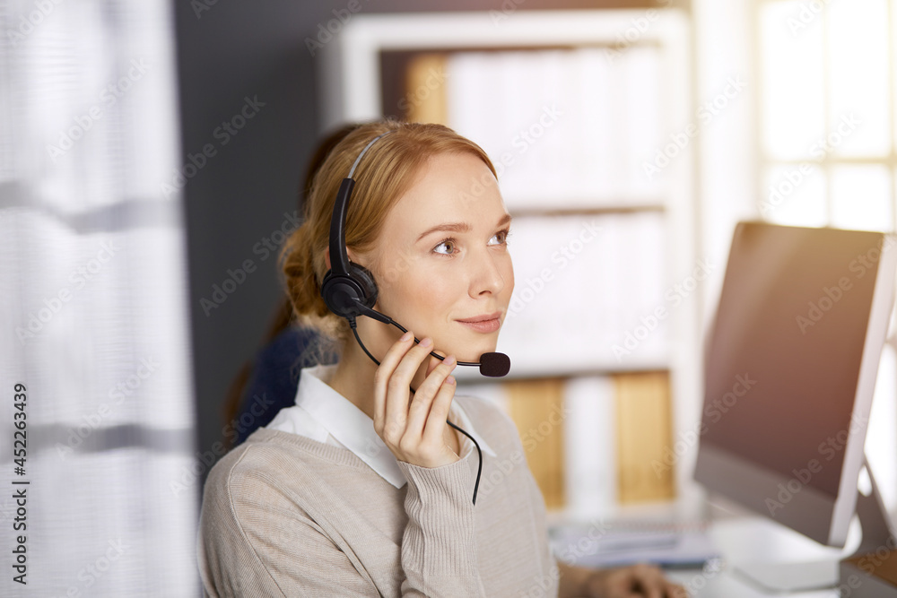 Friendly caucasian businesswoman talking by headset in sunny office. Call center and diverse people group in business Friendly caucasian businesswoman talking by headset in sunny office. Call center and diverse people group in business
