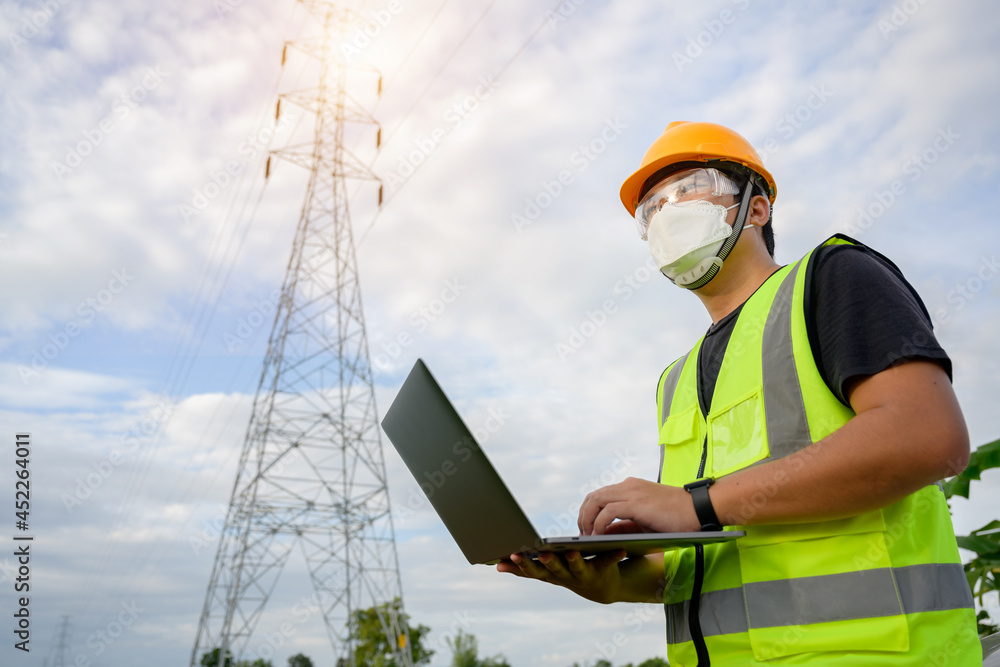 Asian male electrical engineer stands holding a laptop computer at a power plant running and plots to inspect high-voltage poles at a power station. Asian male electrical engineer stands holding a laptop computer at a power plant running and plots to inspect high-voltage poles at a power station.
