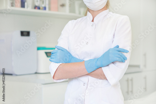 Cropped shot of female doctor cosmetologist in medical gown with arms crossed in medical gloves stands against background of medical office