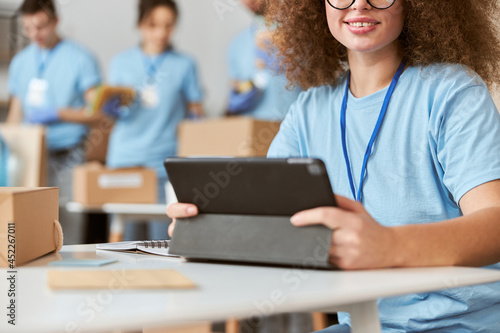 Cropped portrait of young female volunteer in blue uniform using tablet pc and smiling at camera while sitting indoors. Team sorting, packing items in the background © Kostiantyn
