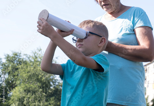 A grandmother with her grandson, a boy, joyfully outdoors on a sunny day, examine the constellations through a toy telescope from a paper tube. 