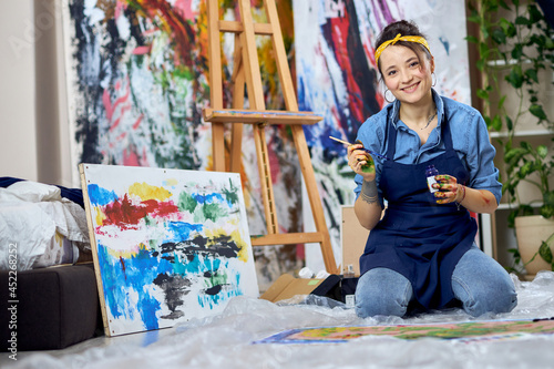 Joyful female painter in apron smiling at camera, holding paintbrush while working on painting, sitting on the floor at home studio workshop