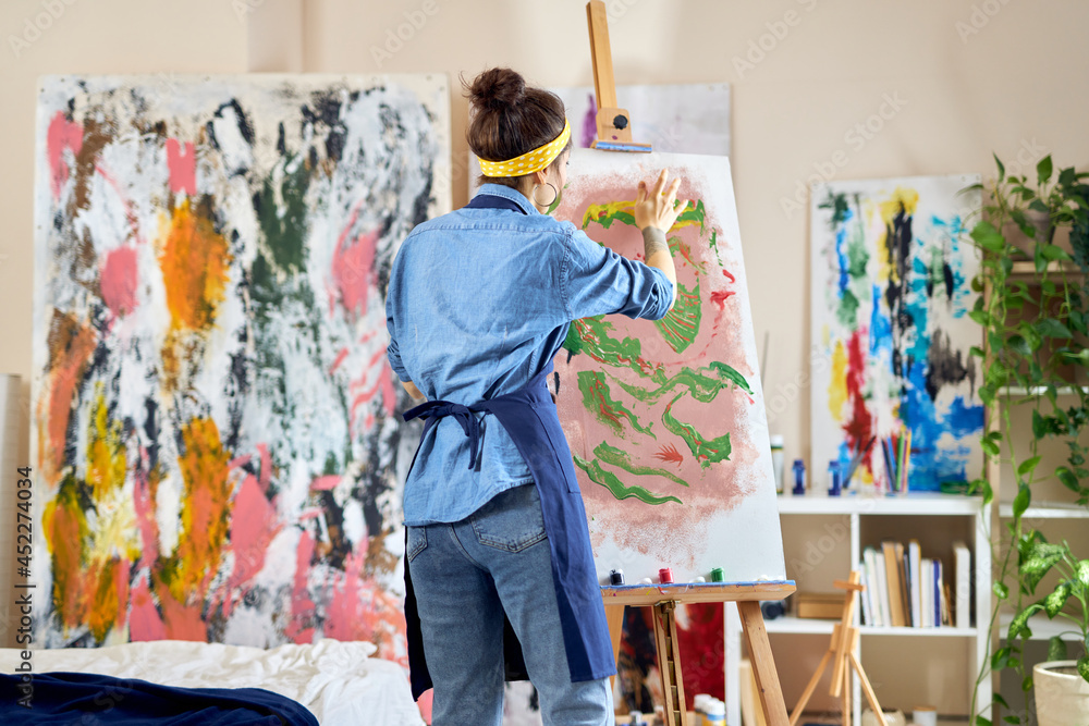 Obraz premium Back view of young female artist in apron creating abstract painting on easel, applying paint on canvas with fingers, standing in living room at home studio