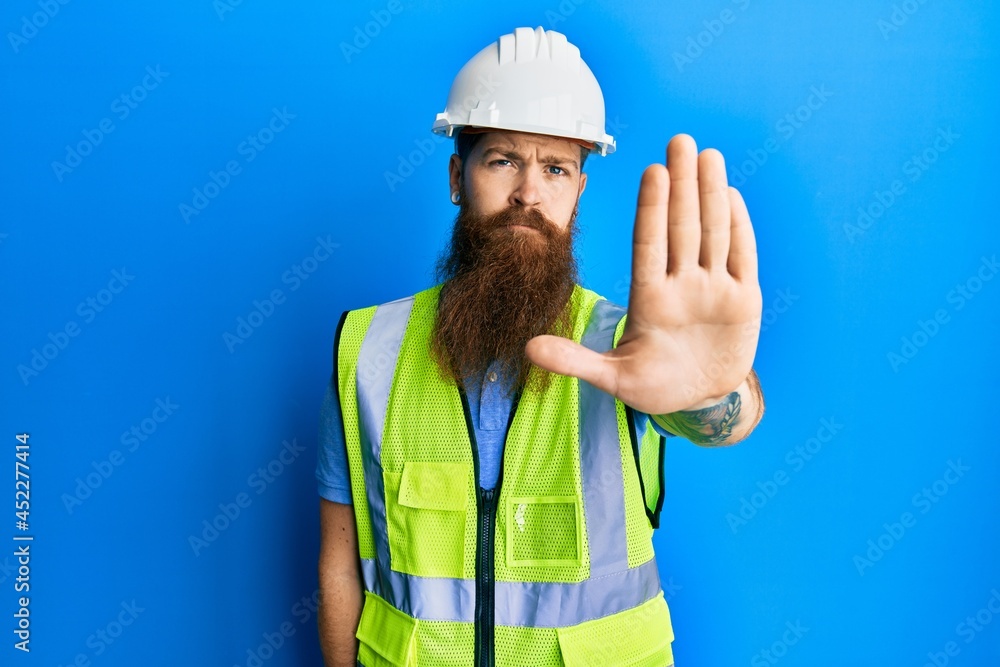 Redhead man with long beard wearing safety helmet and reflective jacket doing stop sing with palm of the hand. warning expression with negative and serious gesture on the face. Redhead man with long beard wearing safety helmet and reflective jacket doing stop sing with palm of the hand. warning expression with negative and serious gesture on the face.