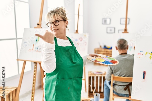 Middle age caucasian woman drawing canvas at art studio pointing thumb up to the side smiling happy with open mouth