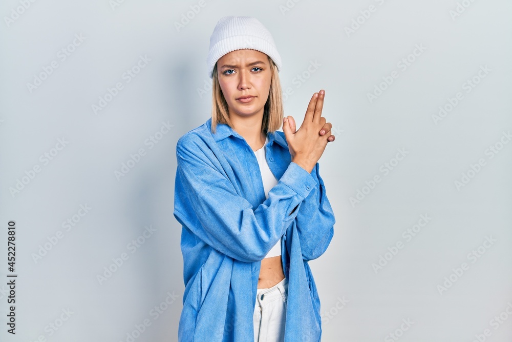 Beautiful blonde woman wearing wool hat holding symbolic gun with hand gesture, playing killing shooting weapons, angry face Beautiful blonde woman wearing wool hat holding symbolic gun with hand gesture, playing killing shooting weapons, angry face
