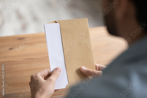 Man sit at table opens envelope with letter or post card inside, close up view over male shoulder. Paper correspondence with information, bank notification, paperwork at workplace, invitation concept photo