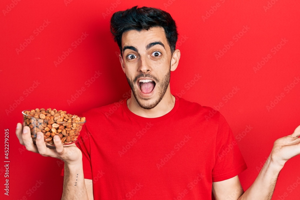 Young hispanic man holding peanuts celebrating achievement with happy smile and winner expression with raised hand Young hispanic man holding peanuts celebrating achievement with happy smile and winner expression with raised hand