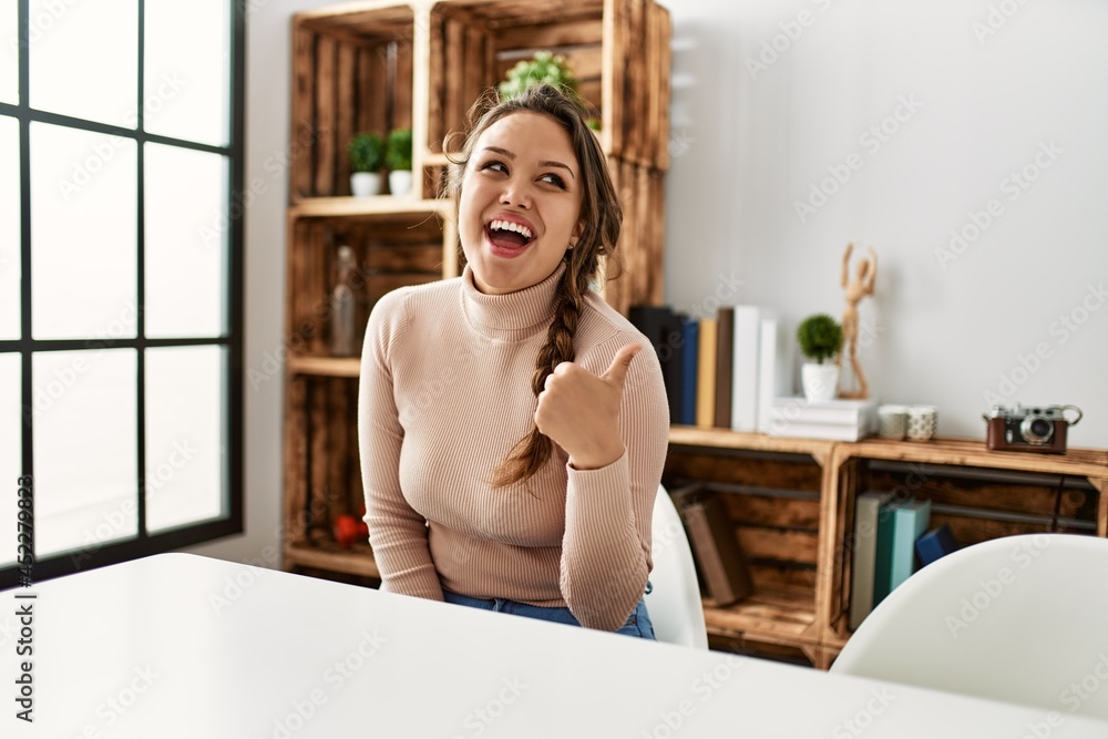 Young hispanic girl wearing casual clothes sitting on the table at home smiling with happy face looking and pointing to the side with thumb up. Young hispanic girl wearing casual clothes sitting on the table at home smiling with happy face looking and pointing to the side with thumb up.