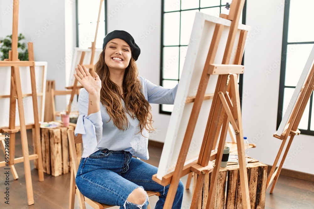 Young hispanic artist woman painting on canvas at art studio waiving saying hello happy and smiling, friendly welcome gesture Young hispanic artist woman painting on canvas at art studio waiving saying hello happy and smiling, friendly welcome gesture