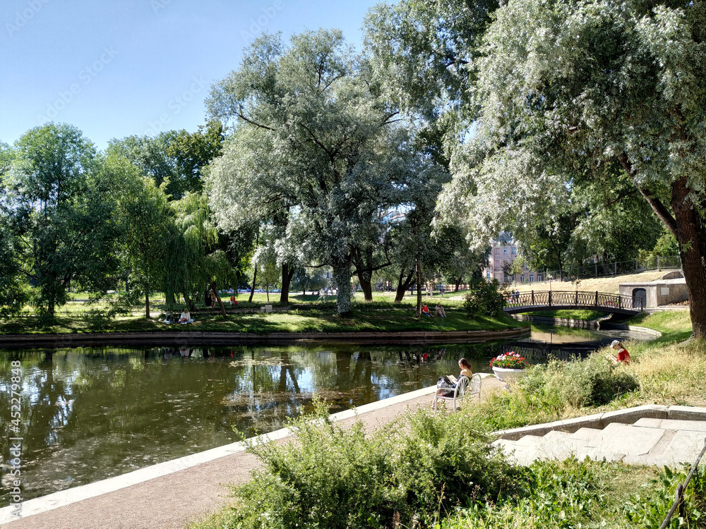Fototapeta premium On a summer day, people relax in the garden. View of the trees, pond, bridge in the Yusupovsky Garden in the old center of St. Petersburg. Recreation places for residents of the city of St. Petersburg