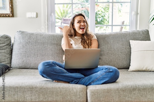 Beautiful young brunette woman sitting on the sofa using computer laptop at home smiling with happy face looking and pointing to the side with thumb up.