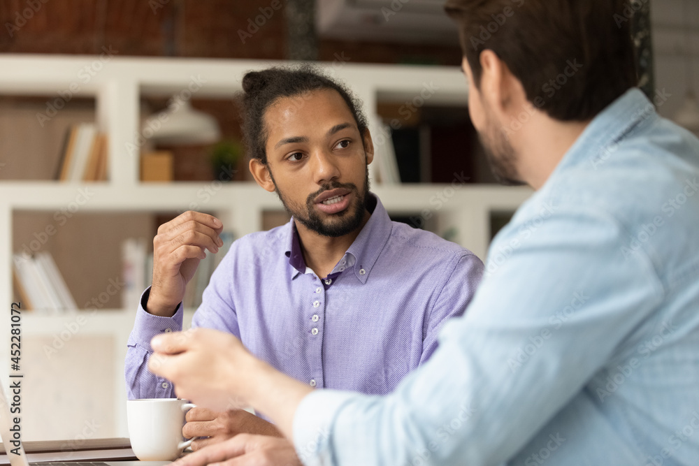 Multi ethnic diverse colleagues discuss collaborative task. African and Caucasian male workmates talking sit at workplace desk share ideas, skills, develop strategy solve business at meeting in office