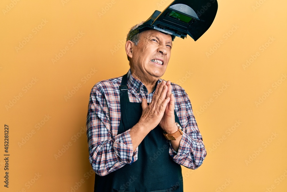 Handsome senior man with grey hair wearing welding protection mask begging and praying with hands together with hope expression on face very emotional and worried. begging. Handsome senior man with grey hair wearing welding protection mask begging and praying with hands together with hope expression on face very emotional and worried. begging.