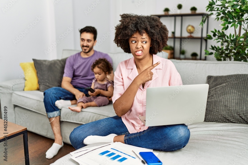 Fototapeta premium Mother of interracial family working using computer laptop at home pointing aside worried and nervous with forefinger, concerned and surprised expression