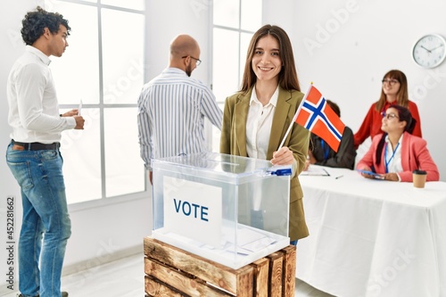 Young norwegian voter woman smiling happy holding norway flag standing by ballot at vote center.